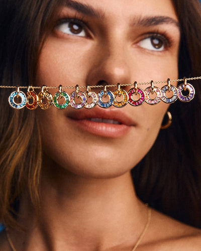 A close-up lifestyle shot of a model wearing gold jewelry, featuring a delicate gold chain draped across her nose with multiple colorful circular ring charms in blue, green, pink, orange, and purple.