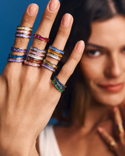 A close-up lifestyle shot of a model's hand wearing multiple colorful gemstone rings, with her face softly blurred in the background against a blue backdrop.