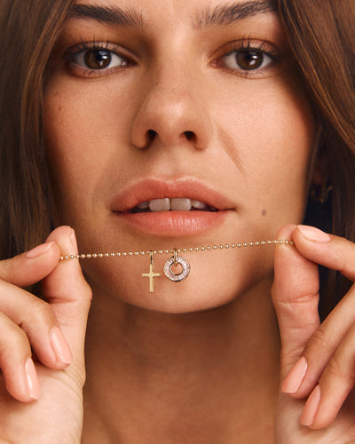 A close-up lifestyle shot of a woman holding a delicate gold chain necklace with a cross pendant and a small circular pendant with a clear stone, against her face.