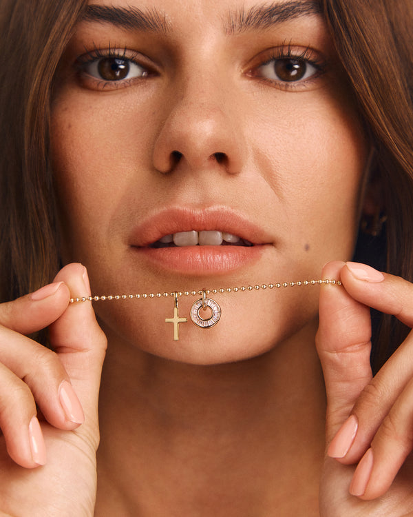 A close-up lifestyle shot of a woman holding a delicate gold chain necklace with a cross pendant and a small circular pendant with a clear stone, against her face.