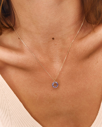 A close-up of a model's neck and chest wearing a delicate gold chain necklace with a small circular pendant featuring a lavender-purple stone.
