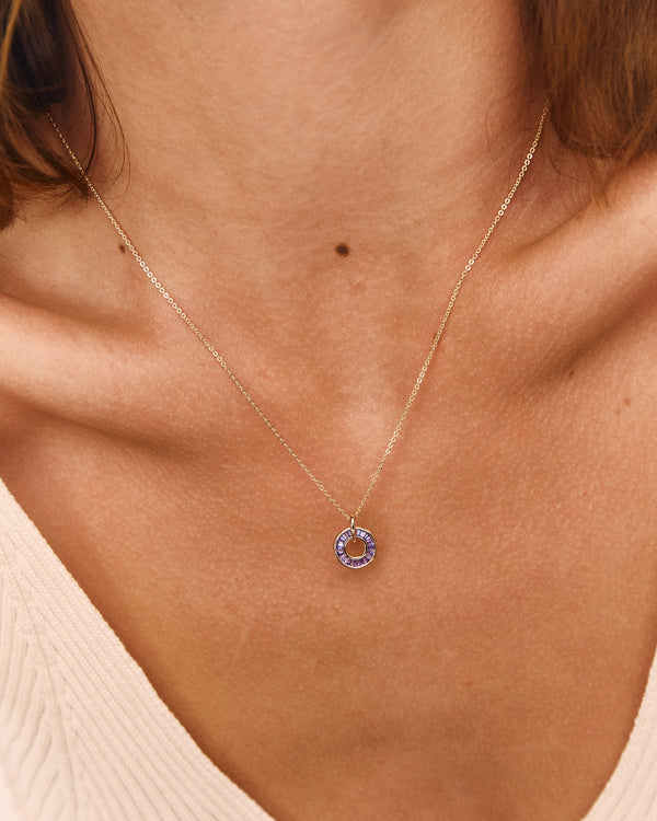 A close-up of a model's neck and chest wearing a delicate gold chain necklace with a small circular pendant featuring a lavender-purple stone.