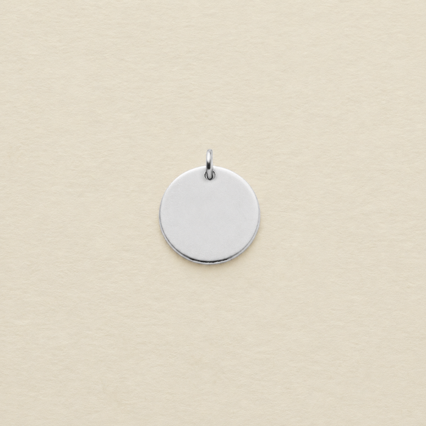 A small round silver disc pendant with a top jump ring, centered on a textured cream-colored background.