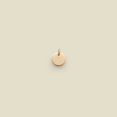 A small, round, gold disc charm with a jump ring at the top, centered on a solid, warm beige background.