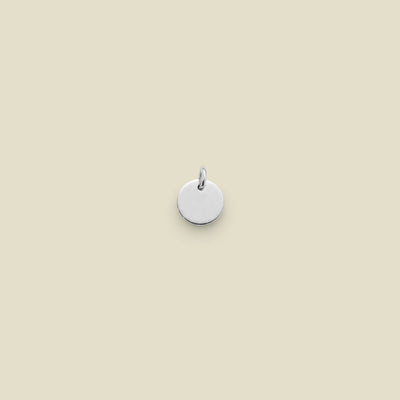 A small, round, silver disc charm with a jump ring at the top, centered on a solid, warm beige background.