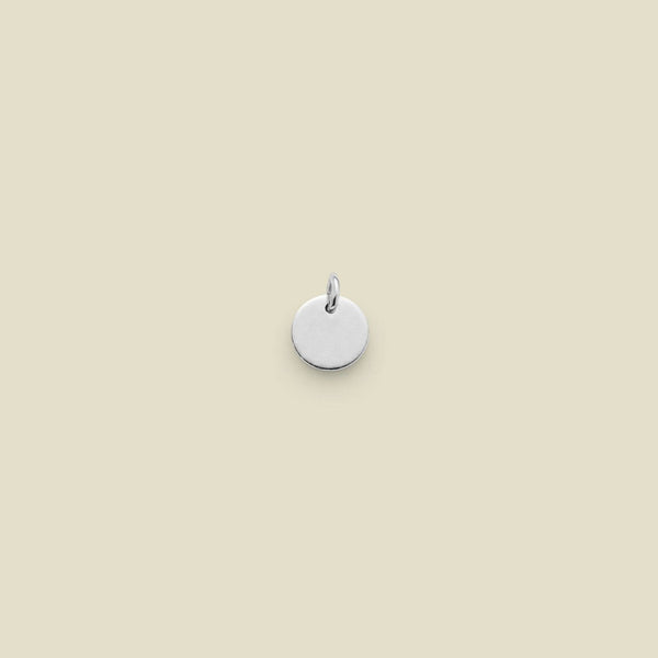 A small, round, silver disc charm with a jump ring at the top, centered on a solid, warm beige background.