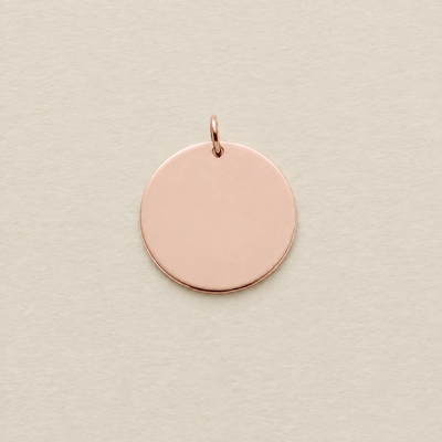A blank, flat circular rose gold disc pendant with a small jump ring attached at the top, shown on a textured light beige background.