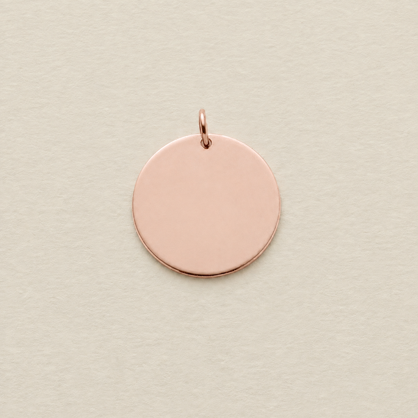 A blank, flat circular rose gold disc pendant with a small jump ring attached at the top, shown on a textured light beige background.