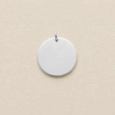 A blank, polished silver round disc pendant with a small jump ring on top, shown on a textured cream-colored background.
