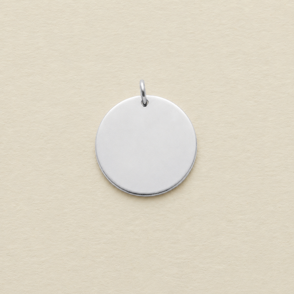 A blank, polished silver round disc pendant with a small jump ring on top, shown on a textured cream-colored background.