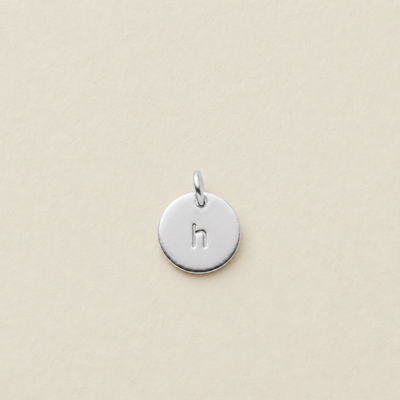 A small round silver disc charm engraved with the lowercase letter 'h' and featuring a top loop, centered on a light beige textured background.