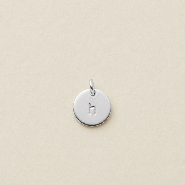 A small round silver disc charm engraved with the lowercase letter 'h' and featuring a top loop, centered on a light beige textured background.