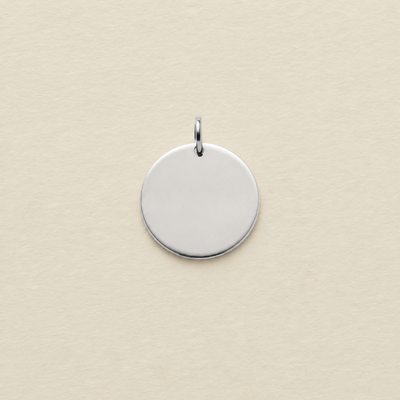 A polished silver round disc pendant with a small top loop, centered on a textured cream-colored background.