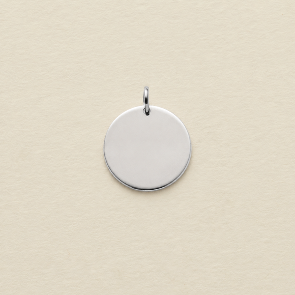 A polished silver round disc pendant with a small top loop, centered on a textured cream-colored background.