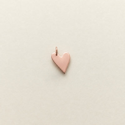 A small polished rose gold heart charm with a top loop, centered on a light beige textured background.