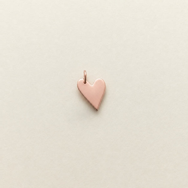 A small polished rose gold heart charm with a top loop, centered on a light beige textured background.