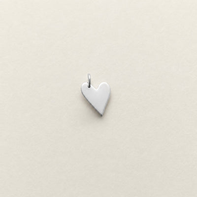 A small, polished silver heart-shaped pendant with a top loop, centered on a textured off-white background.