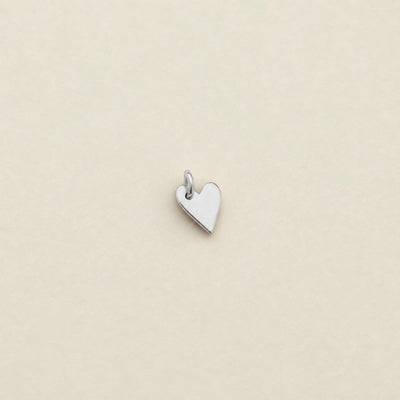 A small polished silver heart charm with a top loop, centered on a plain beige background.