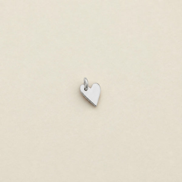 A small polished silver heart charm with a top loop, centered on a plain beige background.
