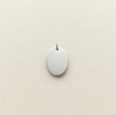A blank, oval-shaped silver pendant with a small top loop, shown on a textured cream-colored background.