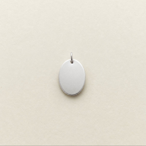 A blank, oval-shaped silver pendant with a small top loop, shown on a textured cream-colored background.