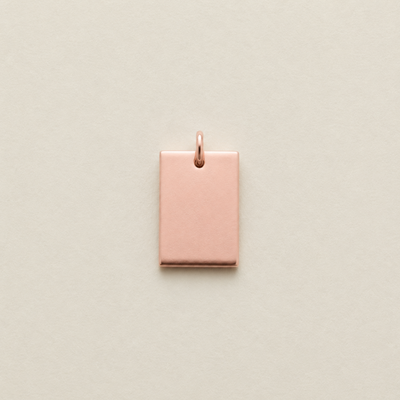 A small rectangular rose gold metal charm with a top loop, centered on a light beige textured background.
