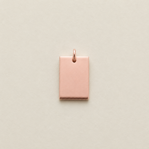 A small rectangular rose gold metal charm with a top loop, centered on a light beige textured background.
