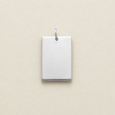 A blank, rectangular silver metal pendant with a small jump ring at the top, shown on a solid beige background.
