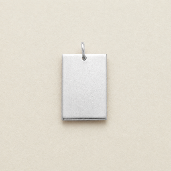 A blank, rectangular silver metal pendant with a small jump ring at the top, shown on a solid beige background.