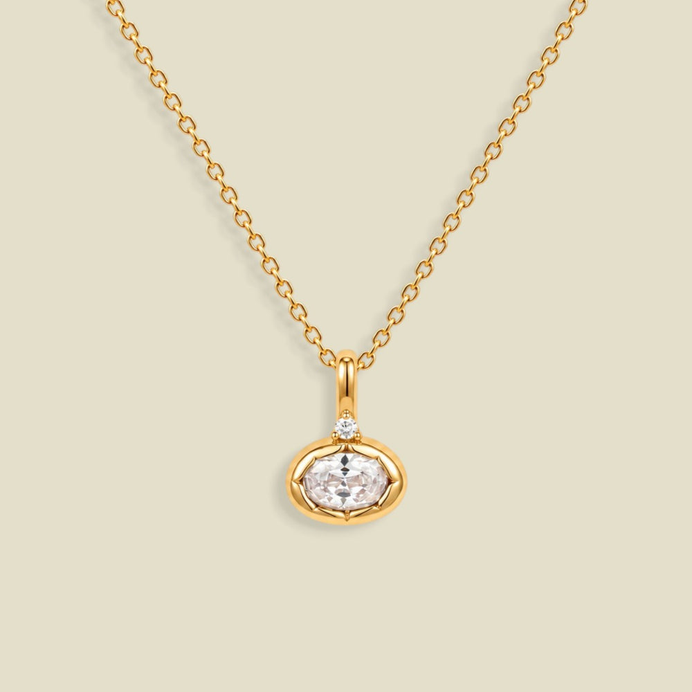 A gold chain necklace with a small oval pendant featuring a clear faceted gemstone and a tiny clear accent stone above it, shown on a light beige background.