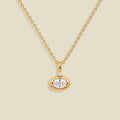 A gold chain necklace with a small oval pendant featuring a clear faceted gemstone and a tiny clear accent stone above it, shown on a light beige background.