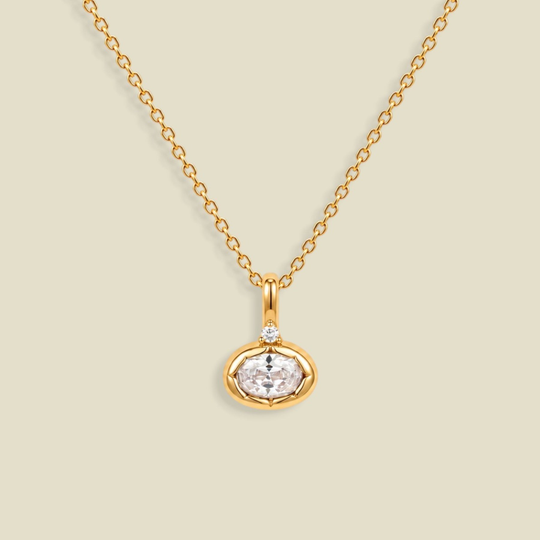 A gold chain necklace with a small oval pendant featuring a clear faceted gemstone and a tiny clear accent stone above it, shown on a light beige background.