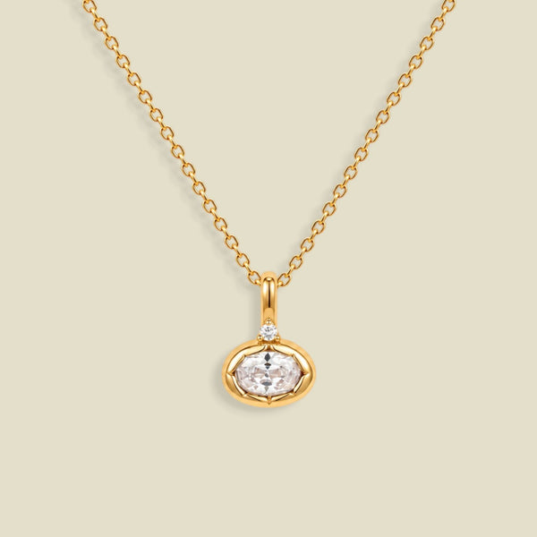 A gold chain necklace with a small oval pendant featuring a clear faceted gemstone and a tiny clear accent stone above it, shown on a light beige background.