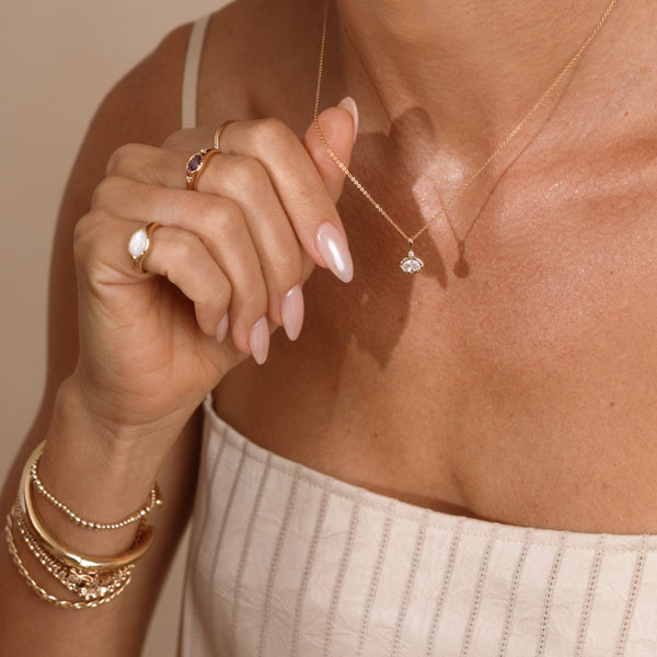 A close-up lifestyle shot of a model wearing a delicate gold chain necklace with a small oval clear crystal pendant, styled with gold rings and bracelets on a neutral beige background.