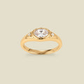 A gold signet ring with a clear center oval bezel-set gemstone, and two small clear accent stones on either side, set against a solid beige background.