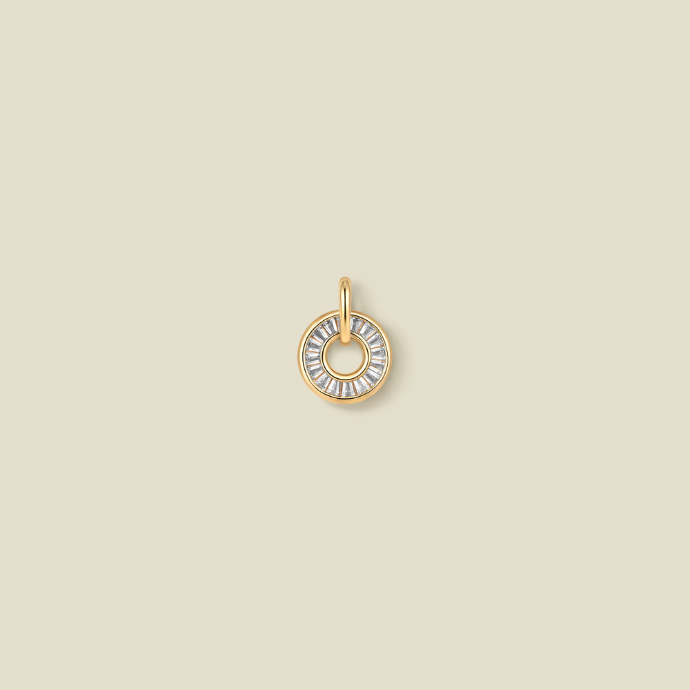 A circular gold pendant featuring clear baguette-cut stones around a hollow center, displayed against a solid light beige background.