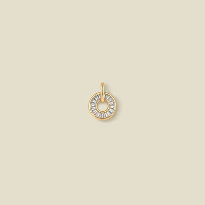 A circular gold pendant featuring clear baguette-cut stones around a hollow center, displayed against a solid light beige background.