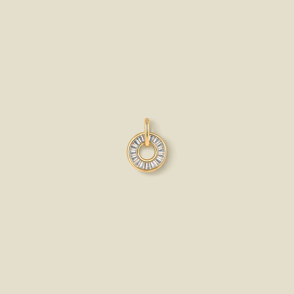 A circular gold pendant featuring clear baguette-cut stones around a hollow center, displayed against a solid light beige background.