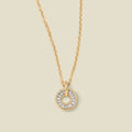 A gold chain necklace with a circular pendant featuring clear baguette-cut stones around a  hollow center, displayed against a solid light beige background.