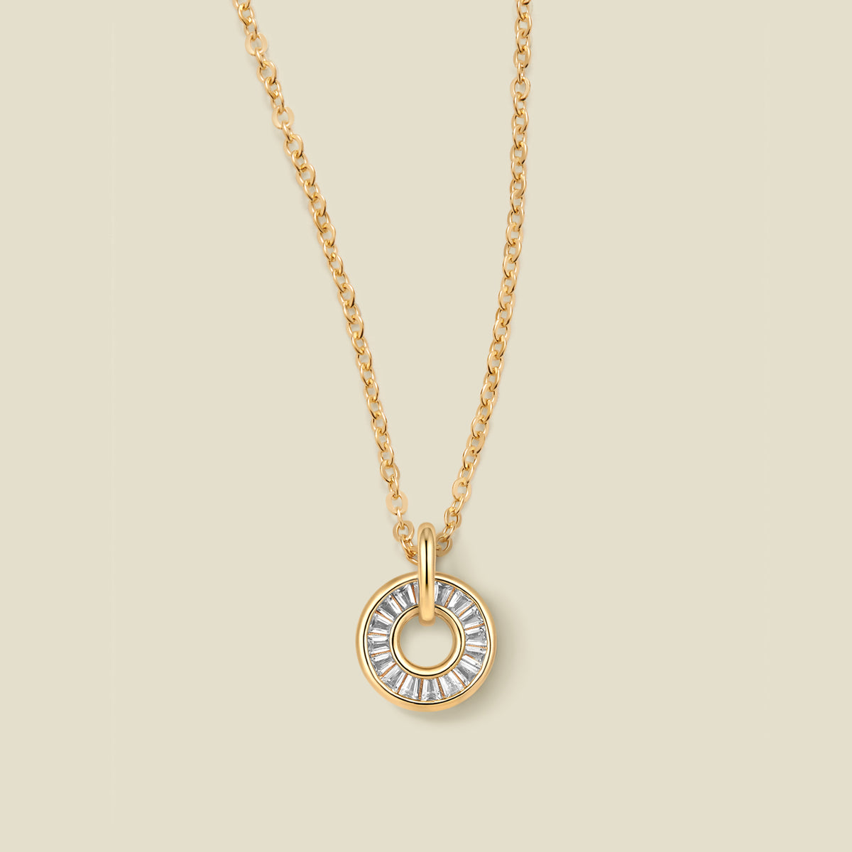 A gold chain necklace with a circular pendant featuring clear baguette-cut stones around a  hollow center, displayed against a solid light beige background.