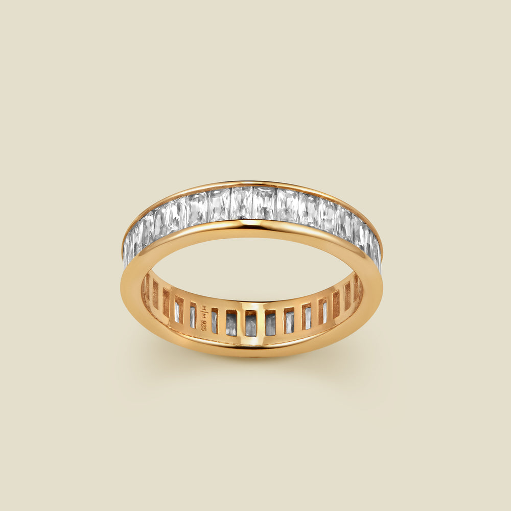 A gold eternity ring featuring a continuous channel setting of baguette-cut clear gemstones, shown on a solid light beige background.