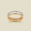A gold eternity ring featuring a continuous channel setting of baguette-cut clear gemstones, shown on a solid light beige background.