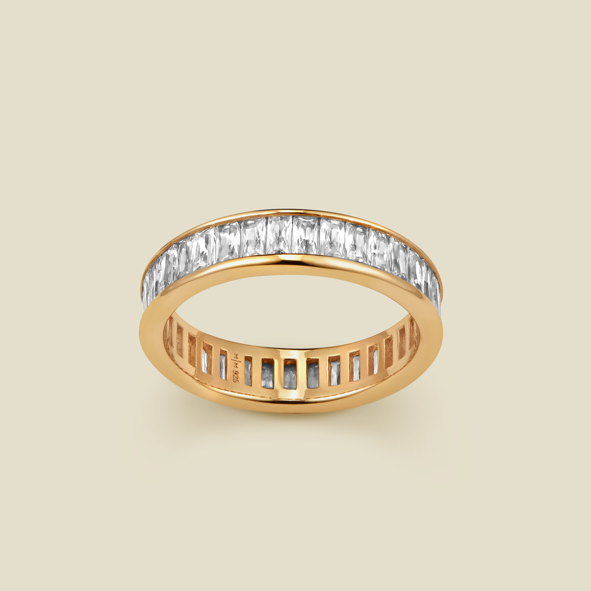A gold eternity ring featuring a continuous channel setting of baguette-cut clear gemstones, shown on a solid light beige background.