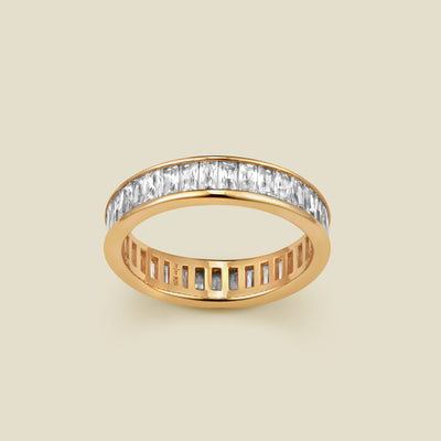 A gold eternity ring featuring a continuous channel setting of baguette-cut clear gemstones, shown on a solid light beige background.