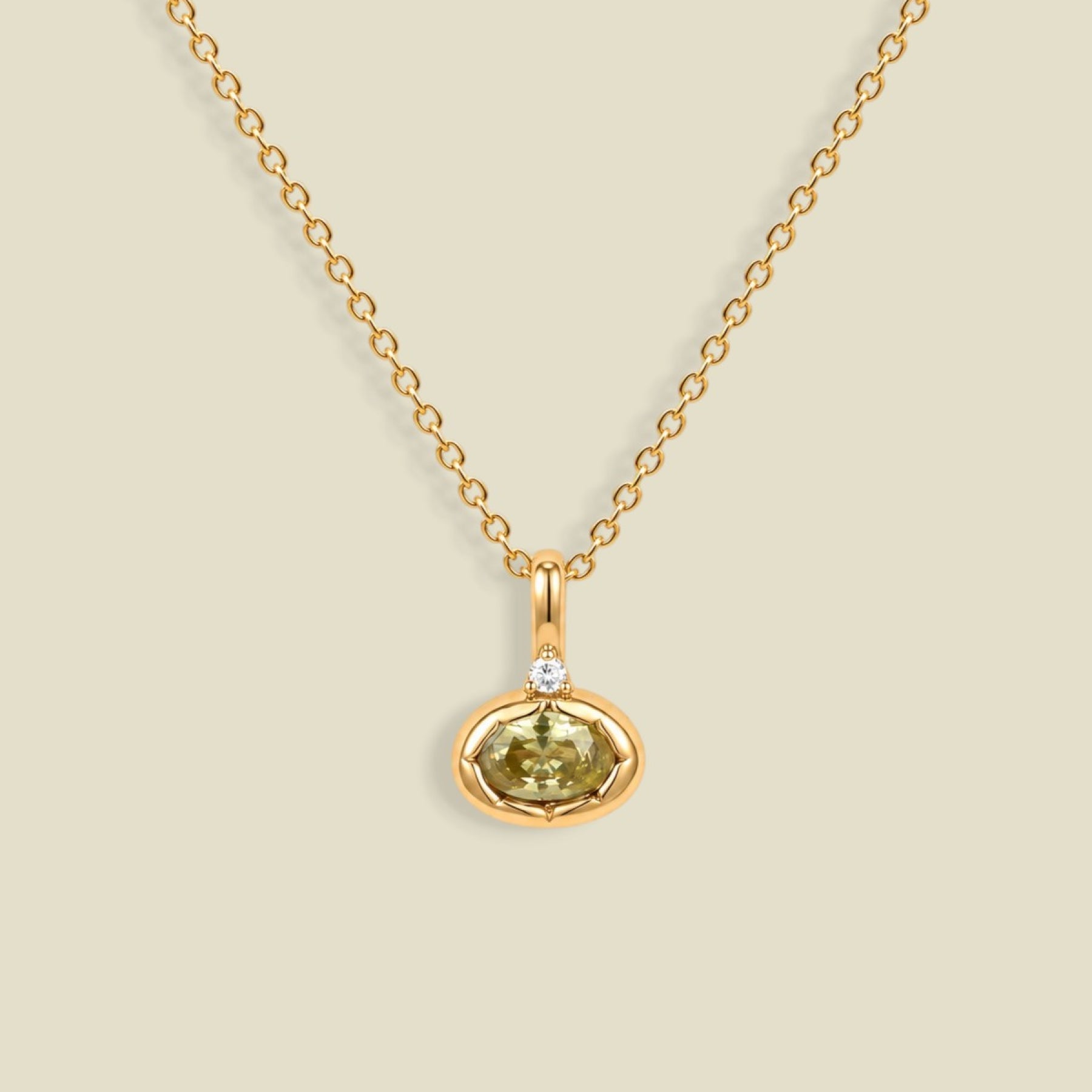A delicate gold chain necklace with a small oval pendant featuring a faceted olive-green gemstone and a tiny clear accent stone above it, displayed on a solid light beige background.