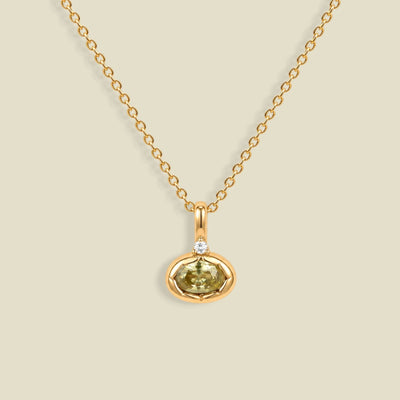 A delicate gold chain necklace with a small oval pendant featuring a faceted olive-green gemstone and a tiny clear accent stone above it, displayed on a solid light beige background.