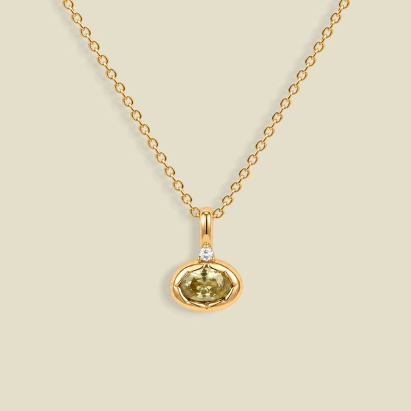 A delicate gold chain necklace with a small oval pendant featuring a faceted olive-green gemstone and a tiny clear accent stone above it, displayed on a solid light beige background.