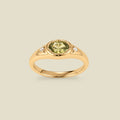 A gold signet ring with a light green center oval bezel-set gemstone, and two small clear accent stones on either side, set against a solid beige background.