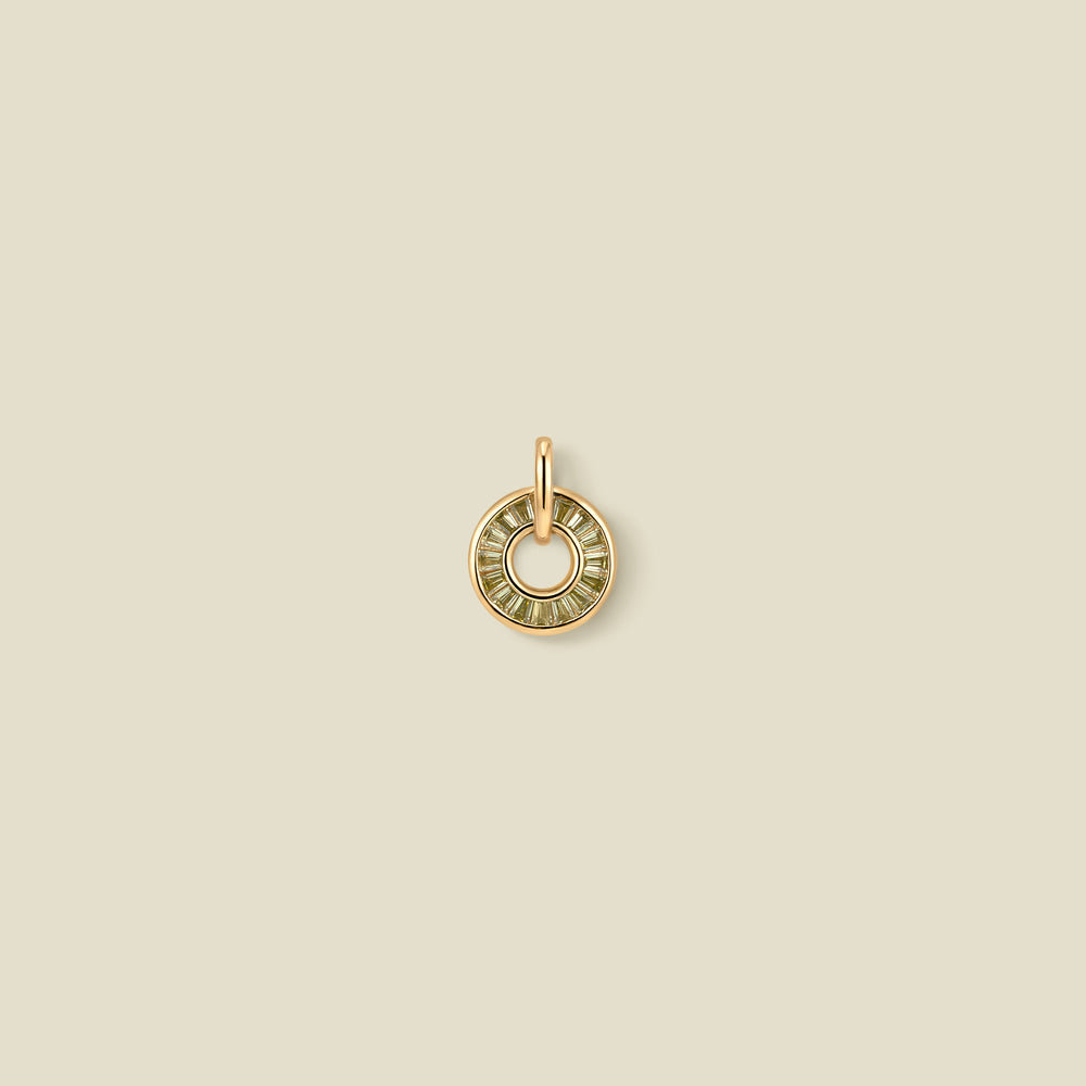 A circular gold pendant featuring light green baguette-cut stones around a hollow center, displayed against a solid light beige background.
