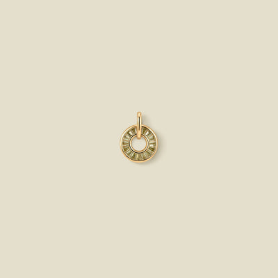 A circular gold pendant featuring light green baguette-cut stones around a hollow center, displayed against a solid light beige background.